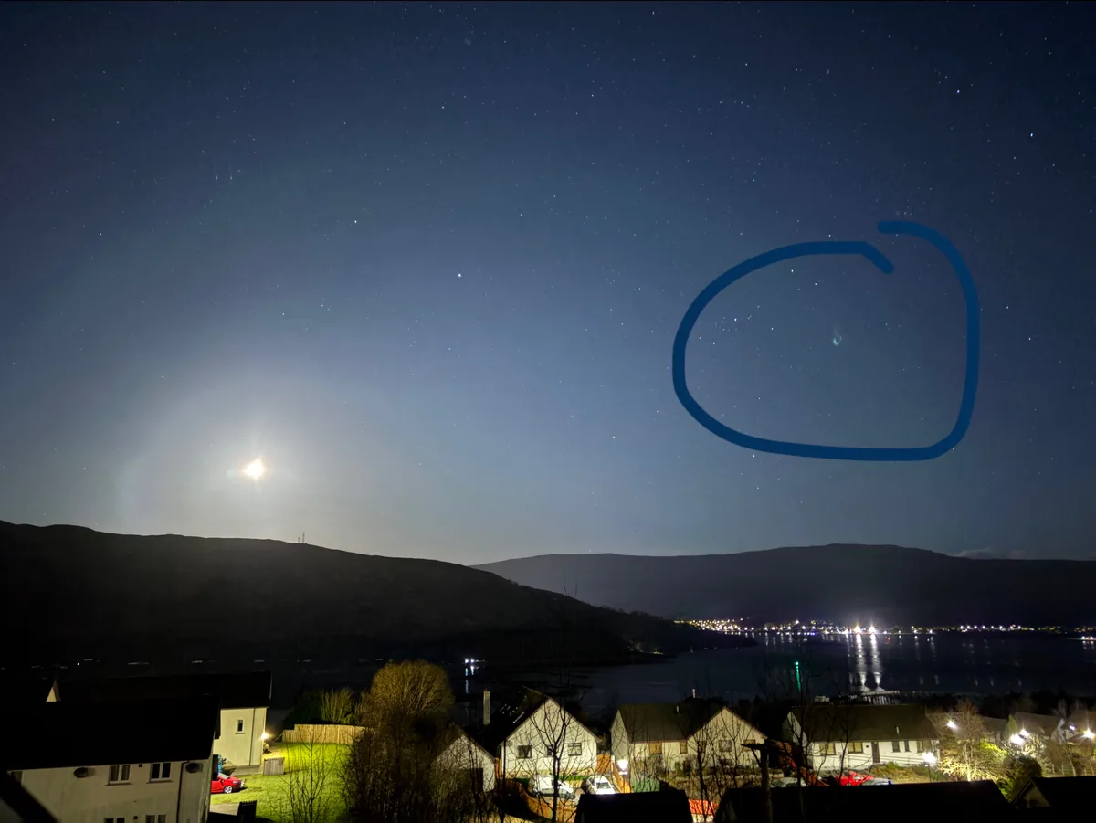 A night sky photo showing stars, a bright moon, and distant hills with houses and water below. A blue hand-drawn circle…
