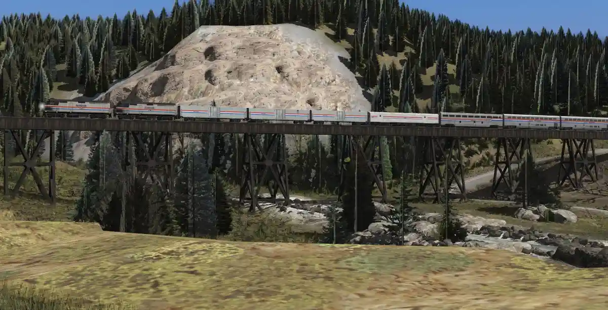 A long Amtrak passenger train travels across a wooden trestle bridge in a mountainous, forested area with a rocky hillside…