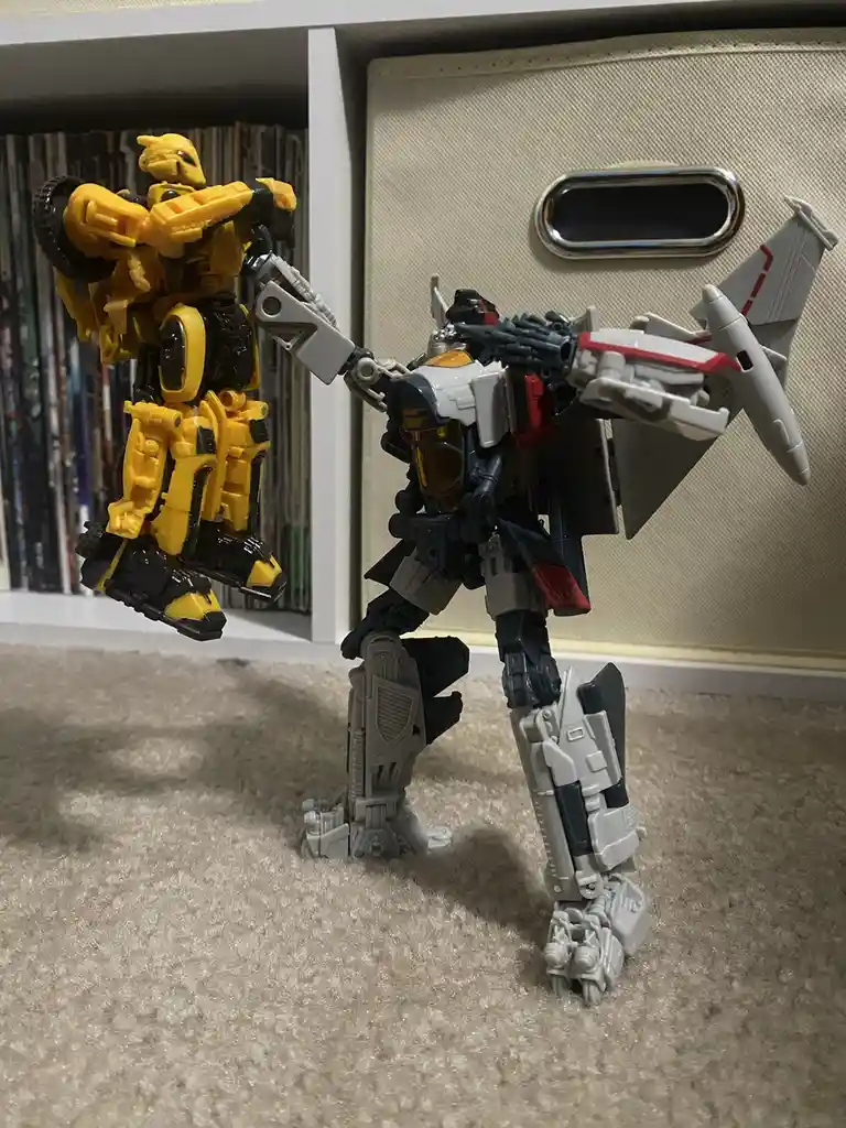 Two transformer action figures are posed on a carpeted floor; a yellow and black figure is being held aloft by a larger…