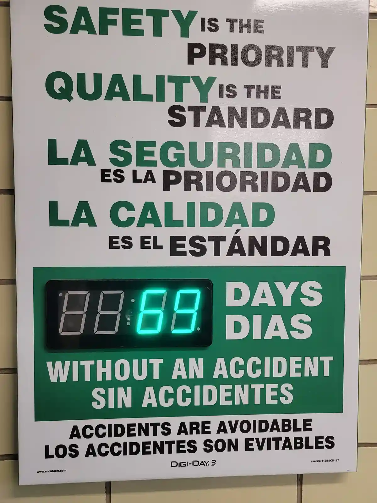 A bilingual safety poster in English and Spanish displays corporate slogans about safety and quality, with a digital counter…