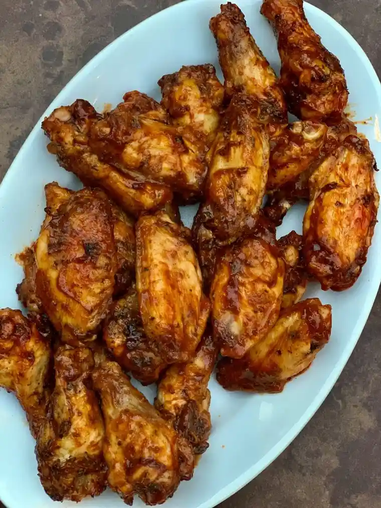An overhead shot of a white oval platter piled high with glossy, sauce-covered chicken wings, resting on a dark countertop…