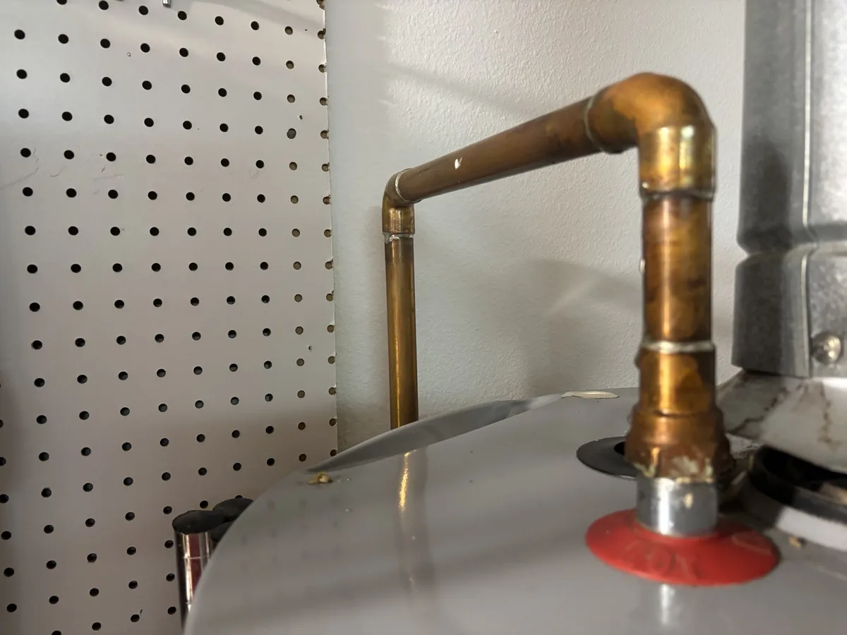 A close-up photo shows a copper pipe connected to a water heater, with a perforated metal panel in the background. The…