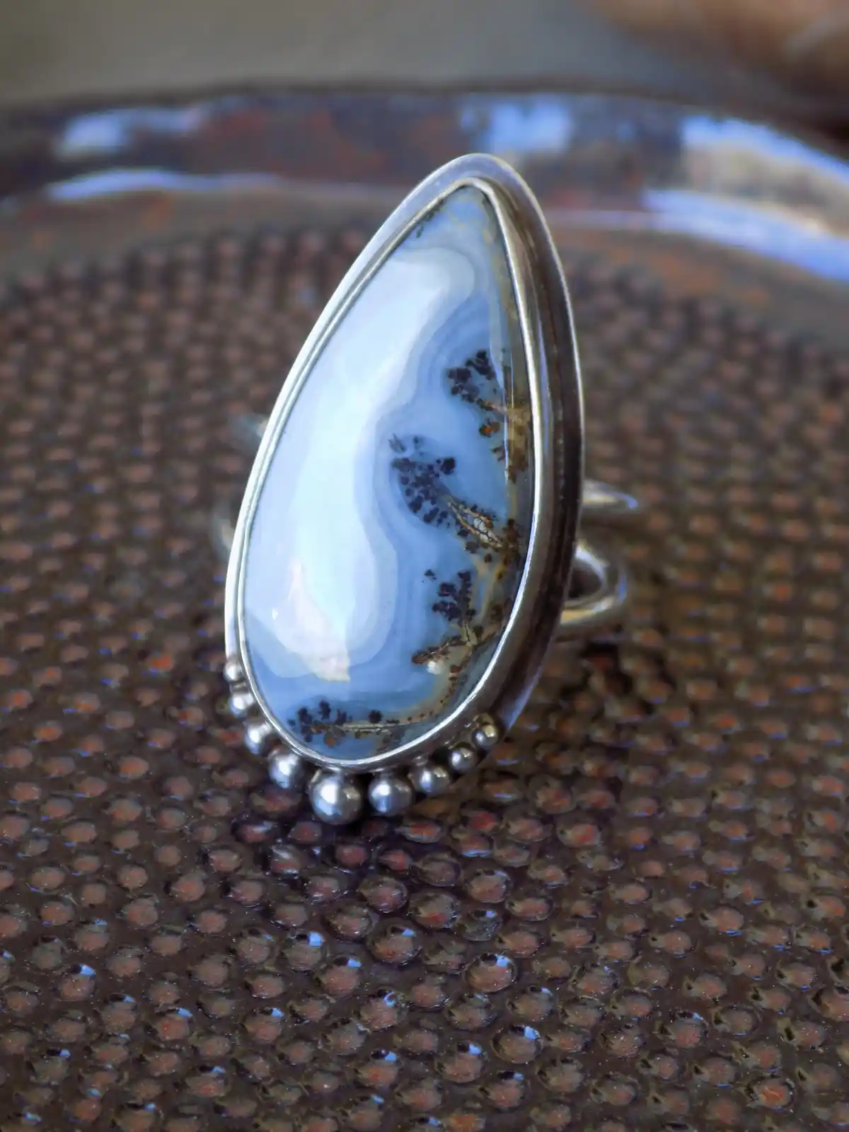 A silver ring with a large, teardrop-shaped blue and white banded stone, possibly dendritic agate, set in a bezel with small…
