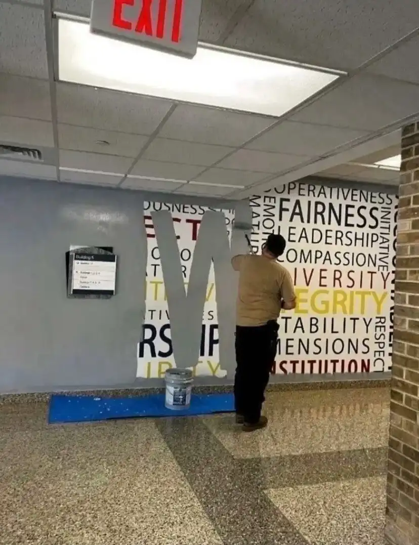 A person is painting a large letter 'M' on a wall in an institutional hallway, covering up words like 'FAIRNESS'…