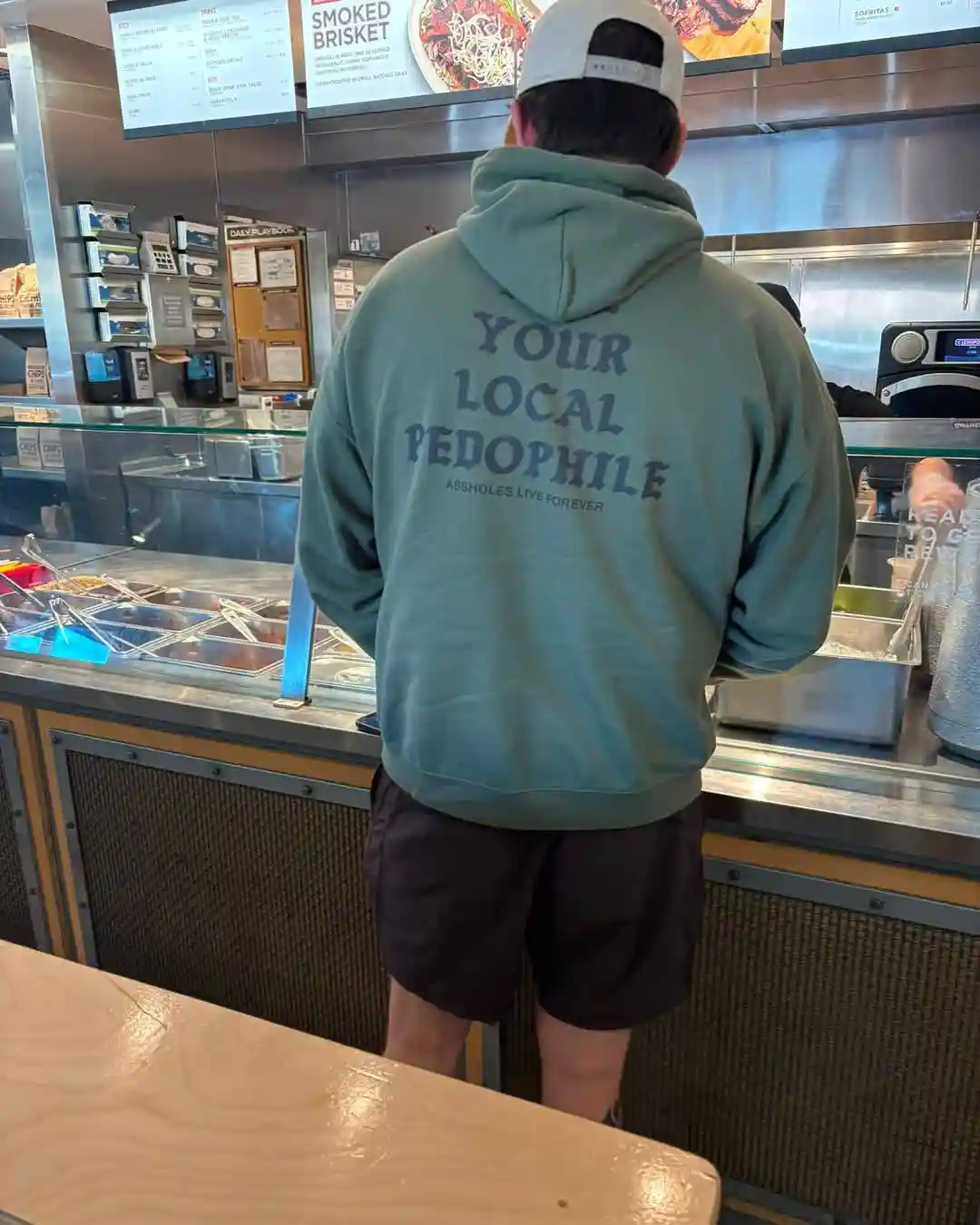 A person viewed from behind, wearing a green hoodie with the text 'YOUR LOCAL PEDOPHILE' and 'ASSHOLES LIVE FOREVER' printed…