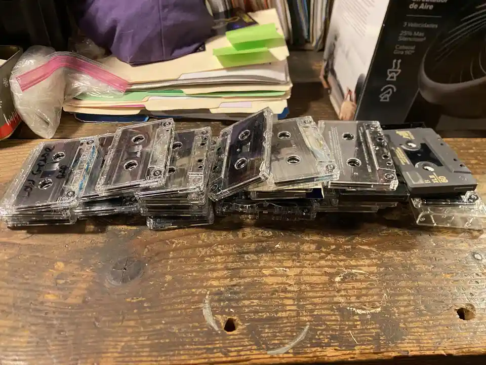 A wooden table holds a long, uneven pile of mostly clear audio cassette tapes, some stacked and others leaning, with one…