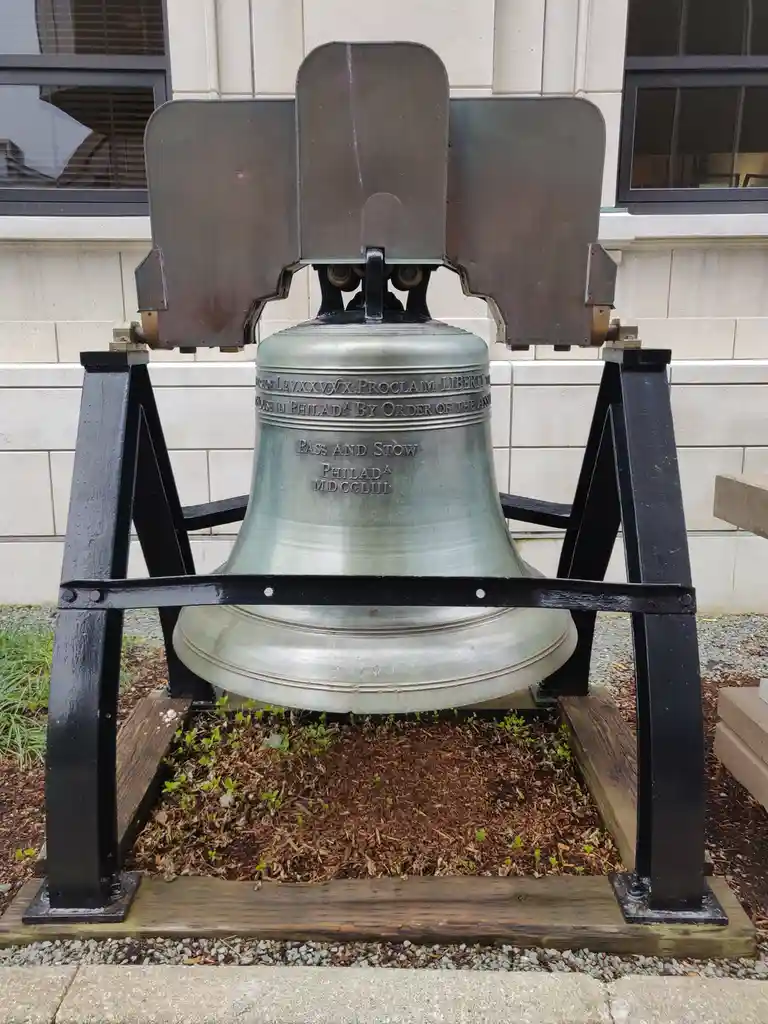 A large, weathered bronze bell mounted on a black metal frame sits outdoors against a light-colored stone building wall. The…