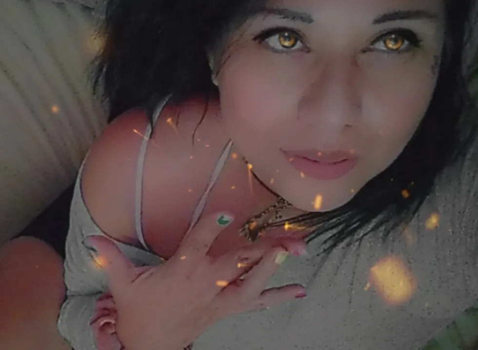 a selfie of a woman with dark hair and golden eyes, wearing a grey tank top and a necklace, looking at the camera with a…