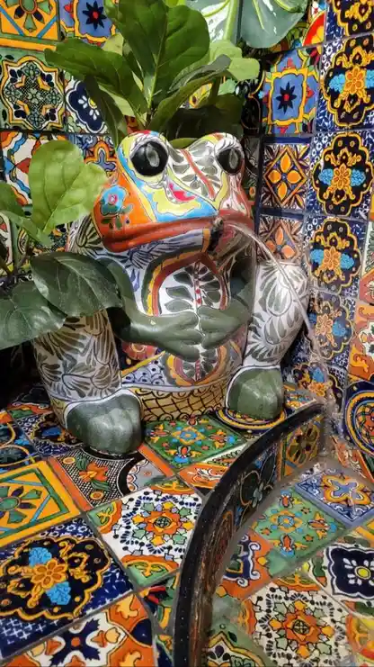 a ceramic frog statue with intricate colorful patterns is positioned as a fountain, water flowing from its mouth into a…