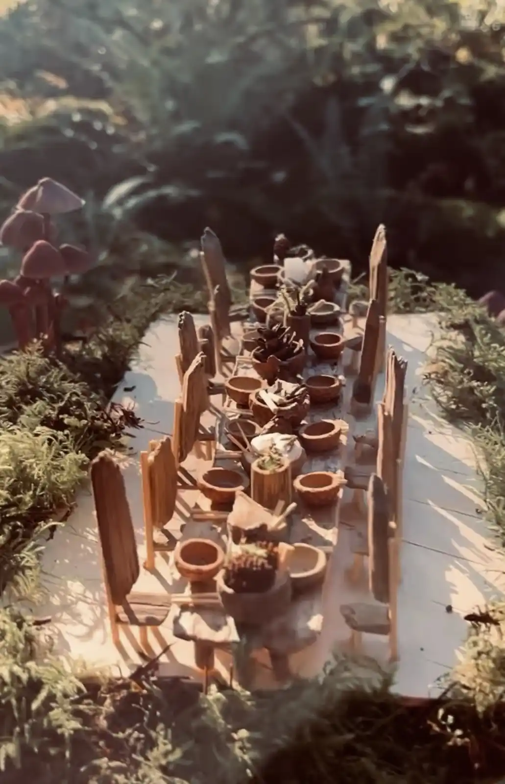 A miniature, rustic wooden dining table set outdoors in a mossy forest, surrounded by tiny chairs and clay bowls filled with…
