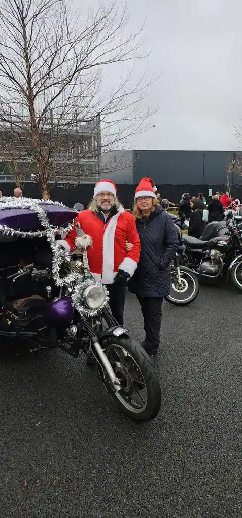 A man and woman, both wearing red Santa hats, stand beside a purple motorcycle decorated with silver tinsel and a gnome. The…