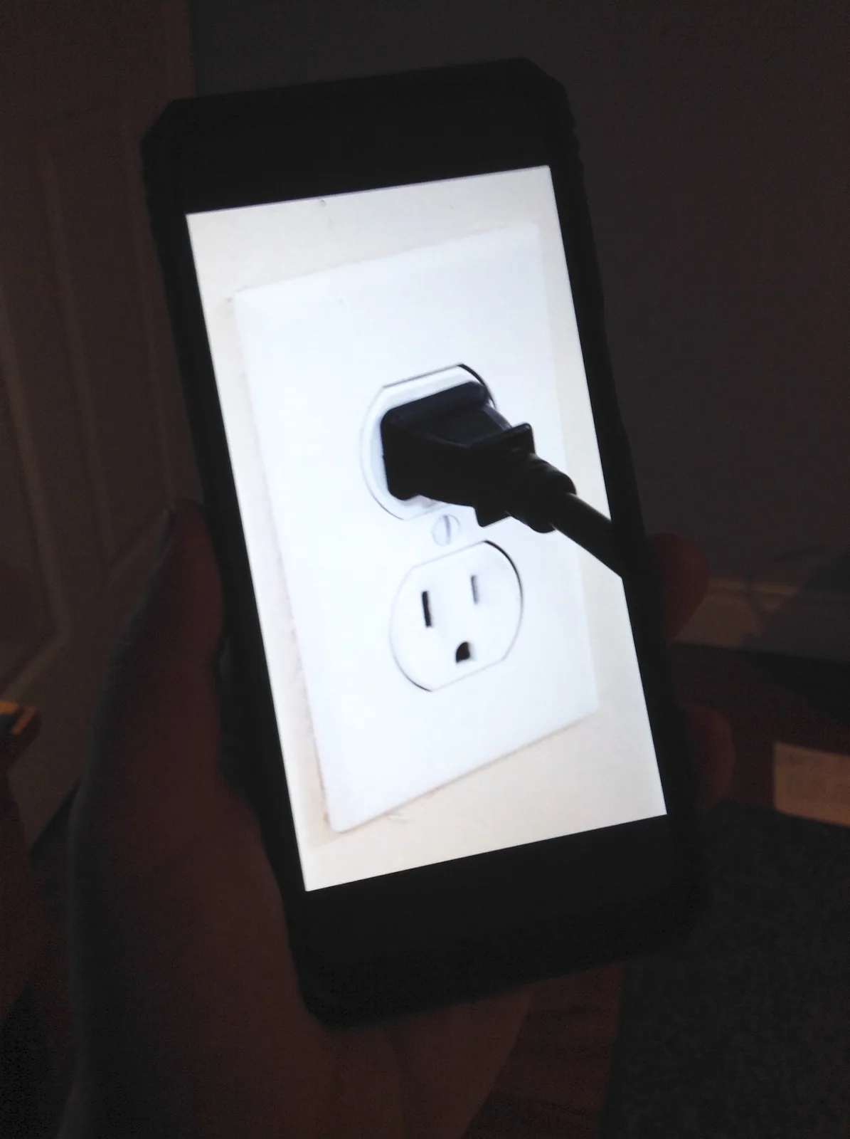 A hand holds a smartphone with a black case, displaying an image of a white wall outlet with a black plug inserted into the…