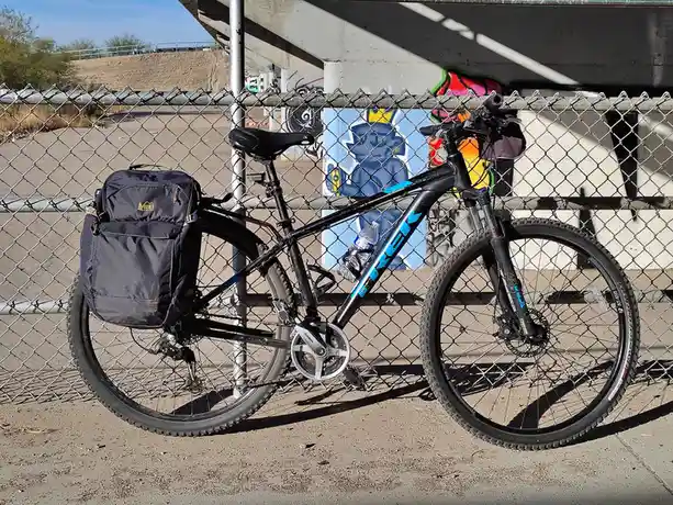 A black mountain bike with a large REI pannier bag on the rear rack and a water bottle in the frame is parked against a…