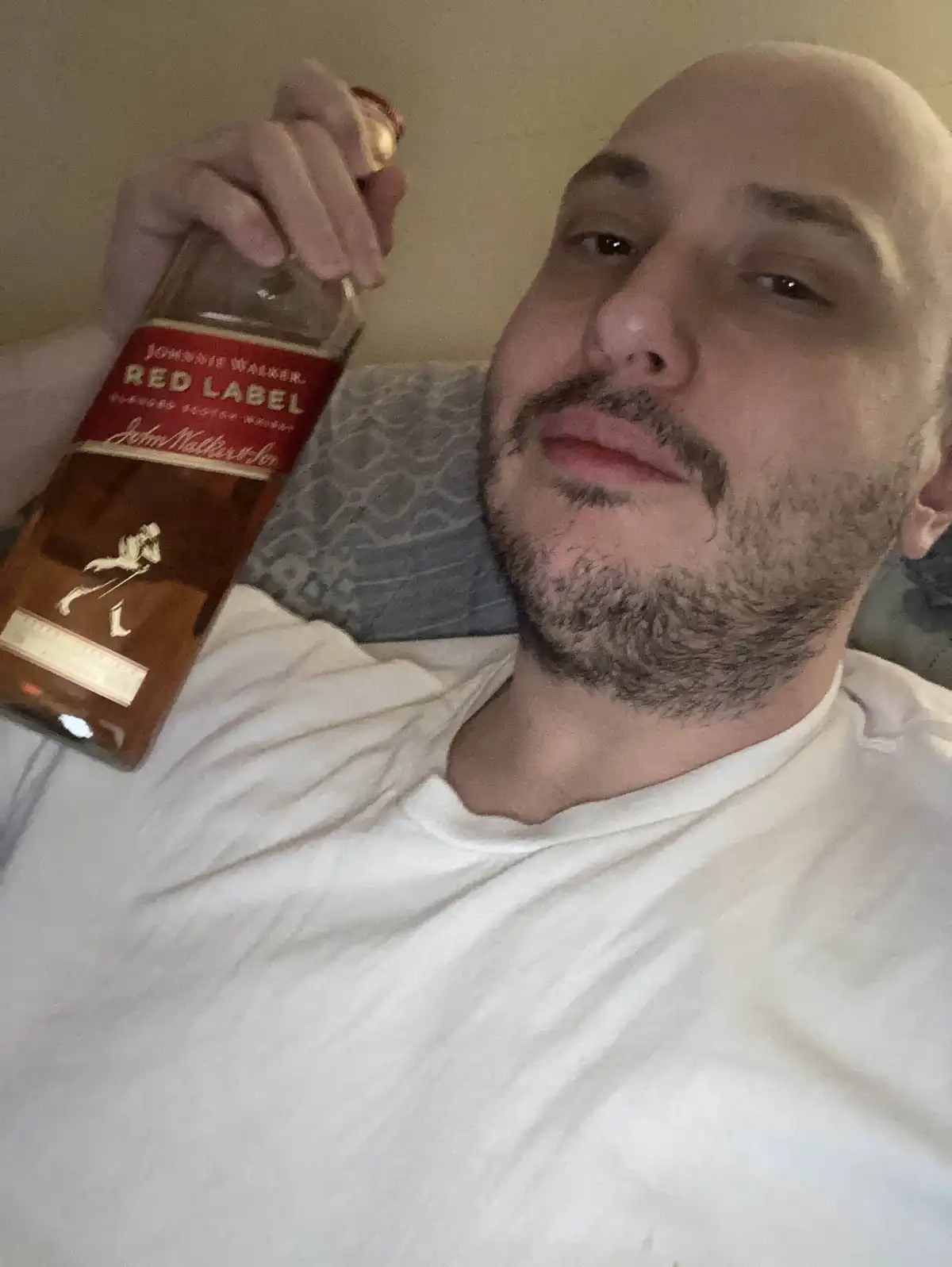 A bald man with a goatee, wearing a white t-shirt, lies on a bed holding a bottle of Johnnie Walker Red Label whisky. He…