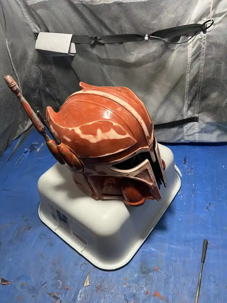 A custom-made Mandalorian-style helmet, freshly painted in a glossy orange and white color scheme, sits atop a white plastic…