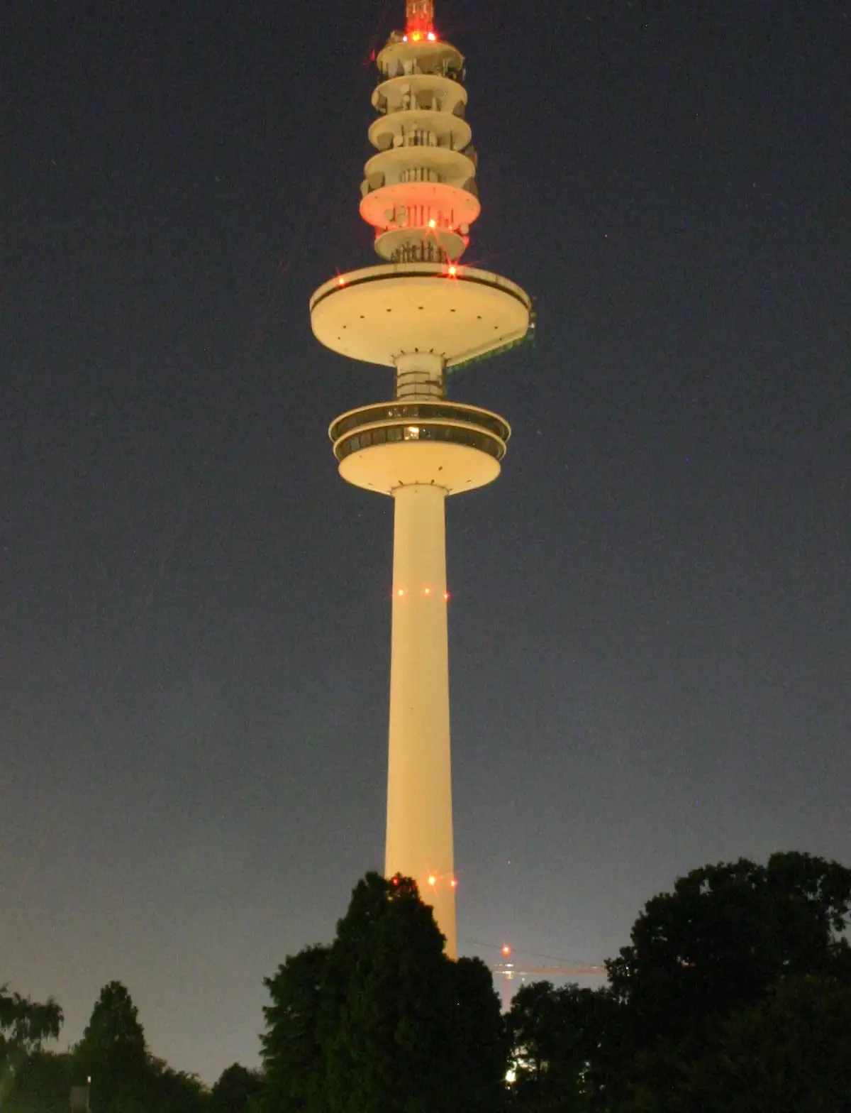 A tall, white, multi-tiered observation tower illuminated at night with red lights, set against a dark sky with trees at the…