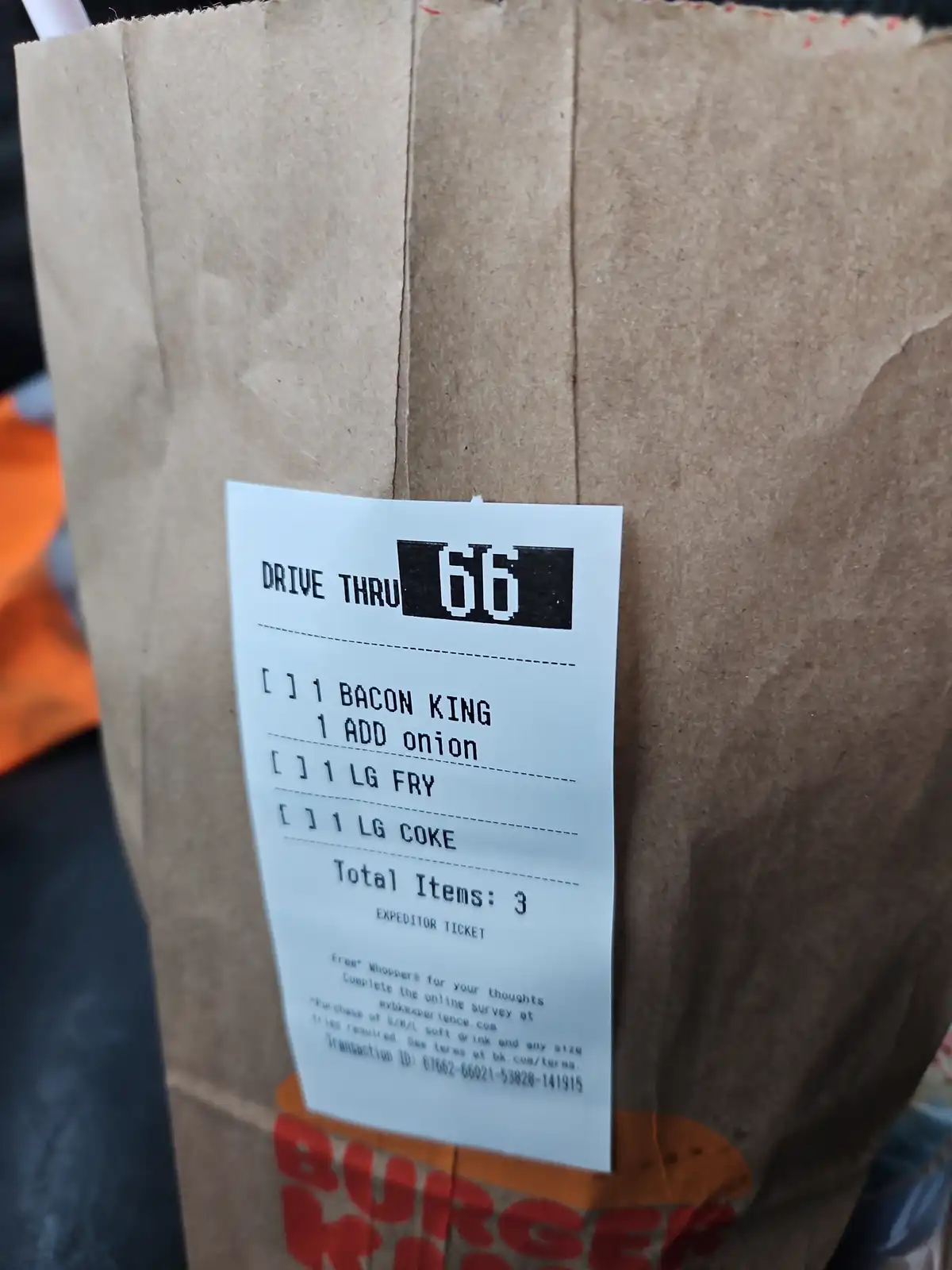 A close-up of a Burger King drive-thru receipt attached to a brown paper bag, showing an order of 1 Bacon King with onion, 1…