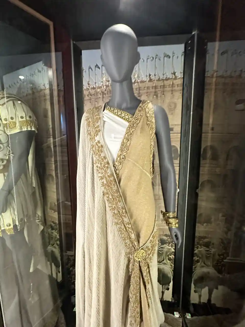 A gray mannequin in a glass display case wears an ornate, gold-embroidered beige toga over a white undergarment, with a gold…