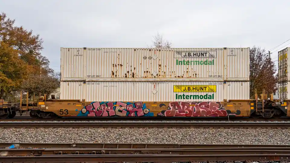 Two stacked J.B. Hunt Intermodal shipping containers sit on a train car covered in graffiti, with the word 'abuse'…
