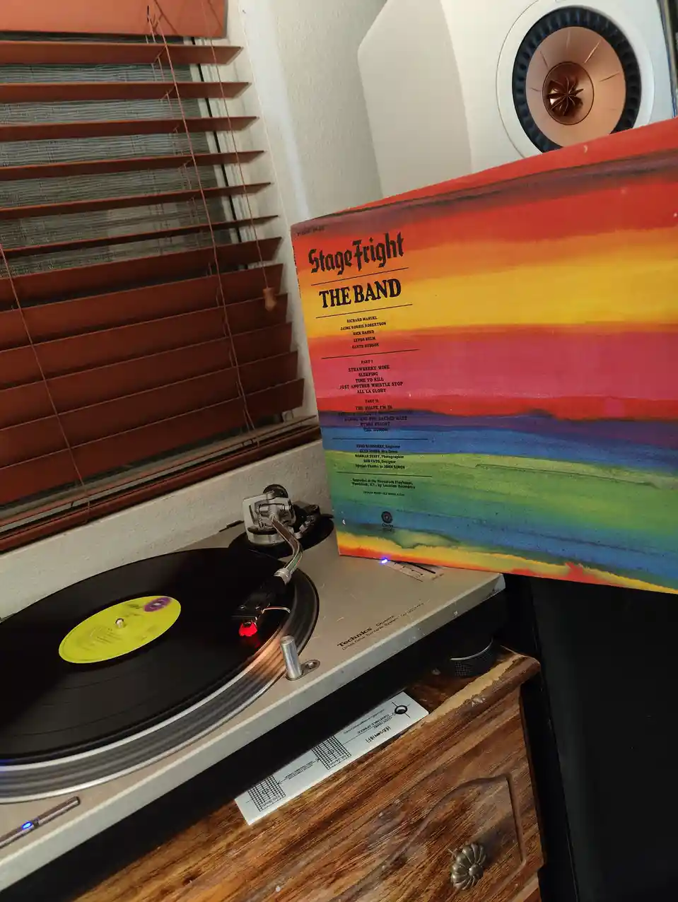 A Technics turntable plays a vinyl record with a yellow label, positioned on a wooden dresser next to a white speaker. The…