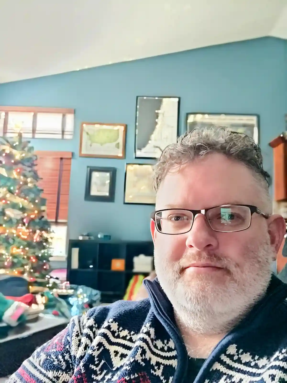 A man with glasses and a gray beard wears a festive patterned sweater while looking at the camera with a deadpan expression…