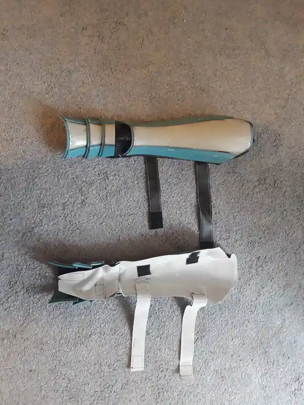 Two prosthetic leg components lie on a carpeted floor; the top one is a finished teal and beige prosthetic with black…