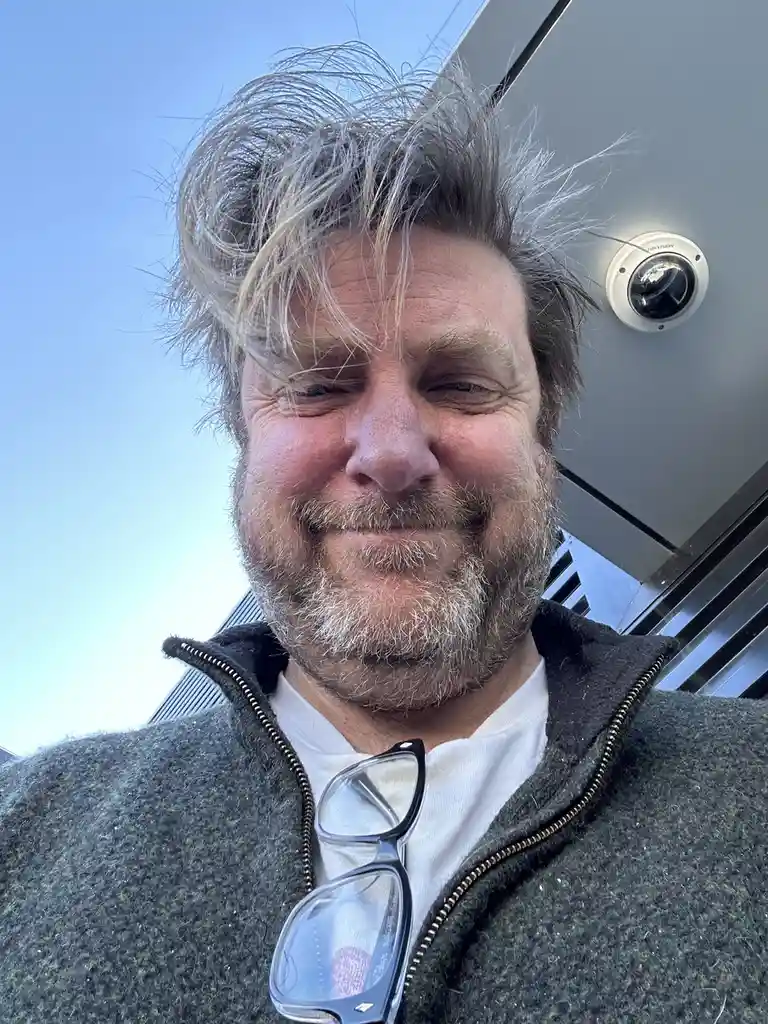 A man with messy gray hair and a beard smiles at the camera while a security camera is visible in the background, implying…