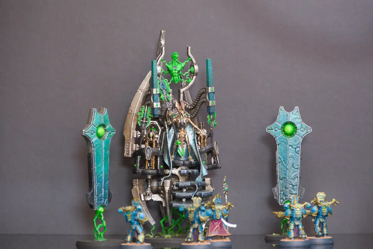 A photo of a Warhammer 40k miniature display featuring a central, heavily detailed green and silver throne with a screaming…