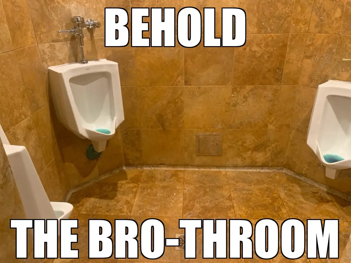A photo of a men's restroom with three white urinals mounted on tan marble tiles, each containing a blue cleaning brush…