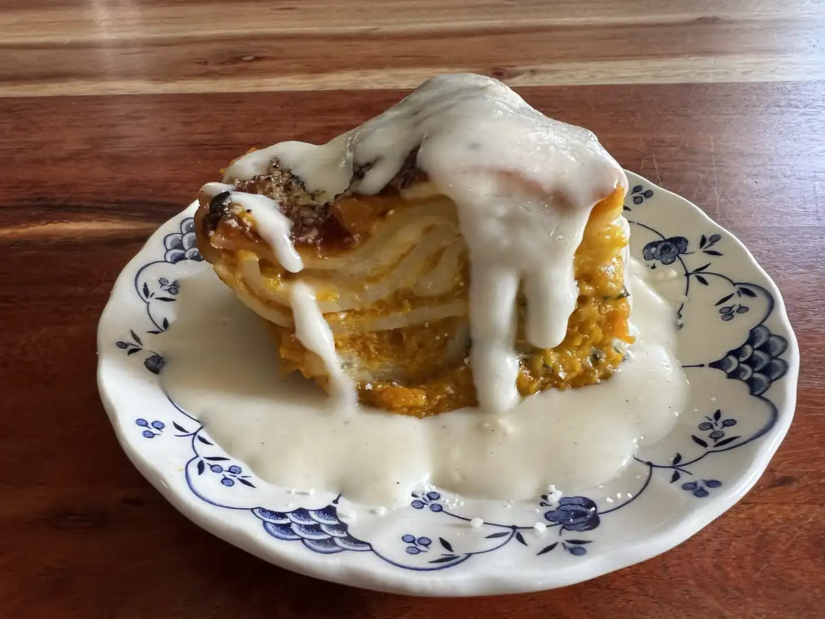 A slice of layered pumpkin pie sits on a white plate with blue floral patterns, heavily drenched in a thick, white gravy…