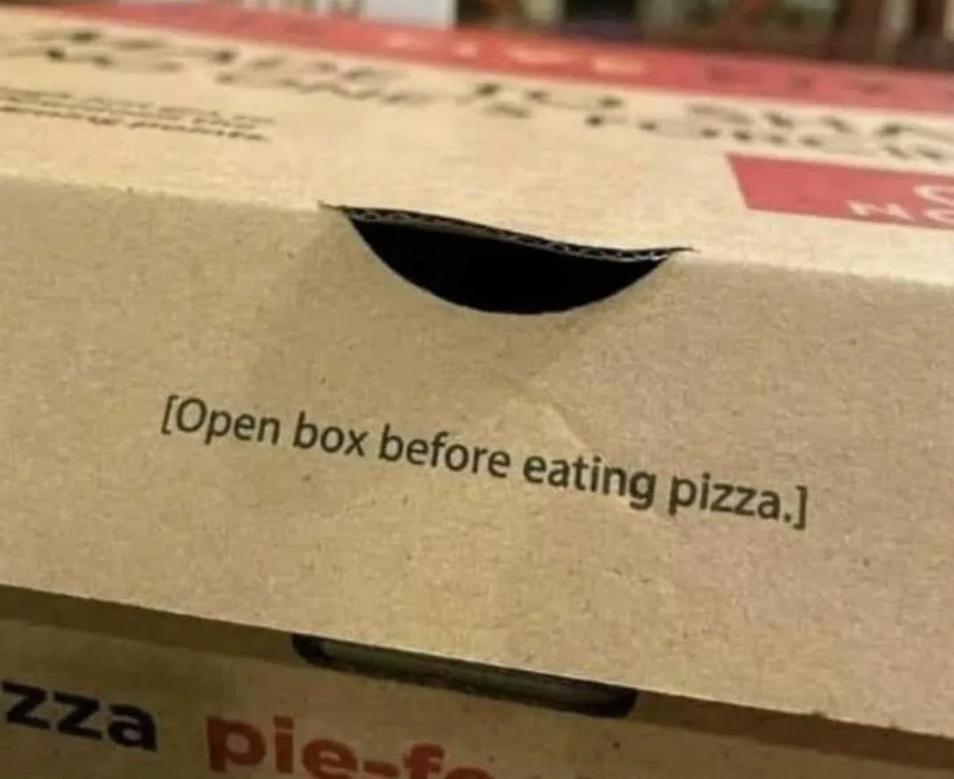 A close-up photo of a cardboard pizza box lid with a small, curved slit cut into it, resembling a mouth. Printed below the…