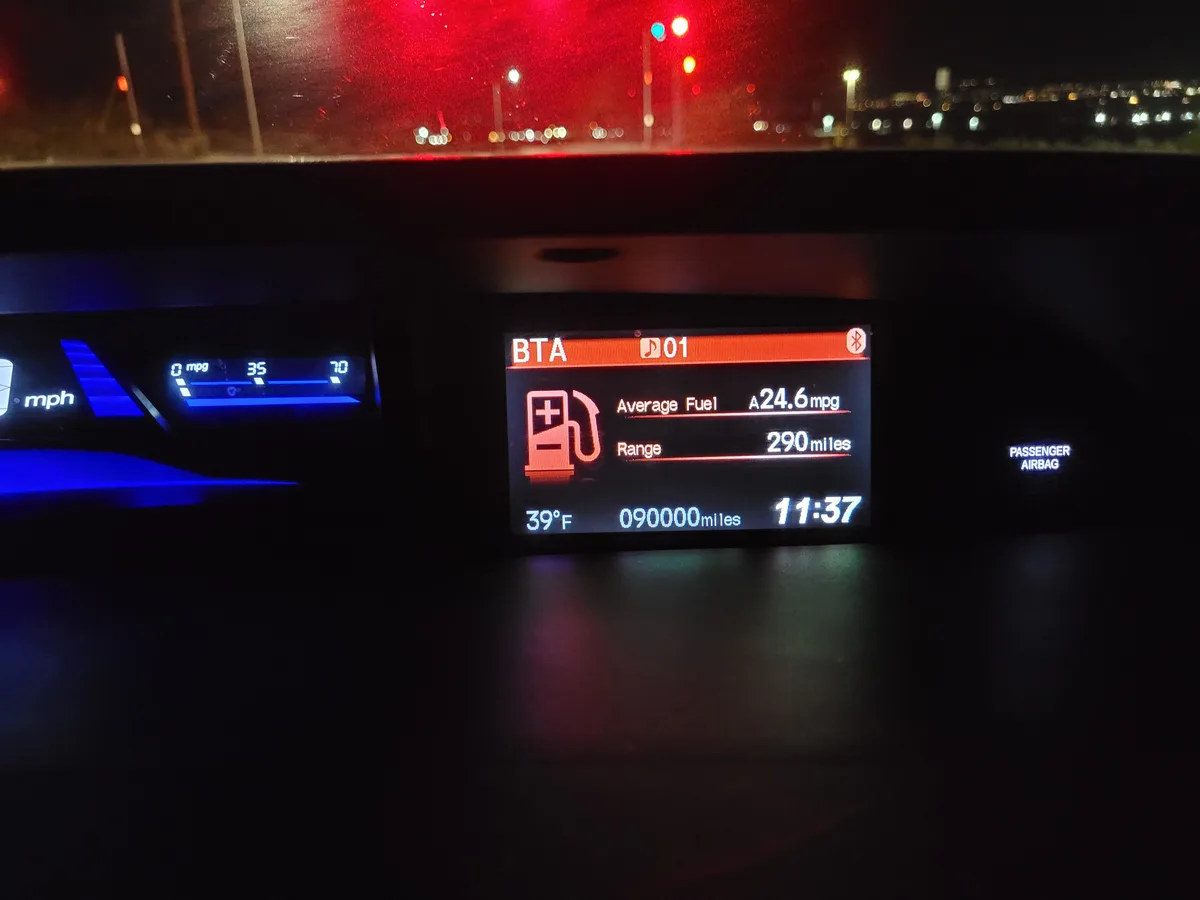 A nighttime car dashboard view showing a digital display with fuel economy stats: average fuel efficiency of 24.6 mpg and a…