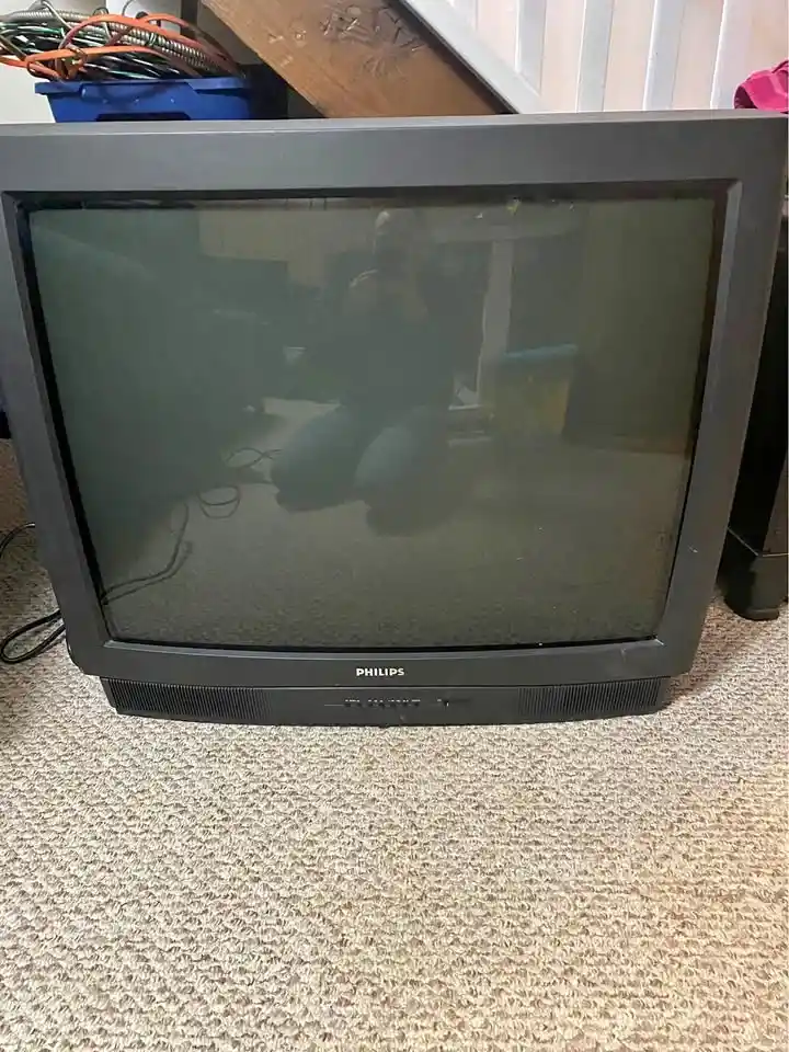 A black Philips CRT television sits on a beige carpet, its screen off and reflecting a person kneeling and taking the photo…