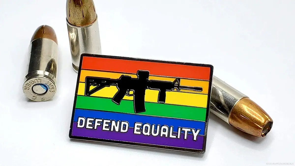 A rectangular pin featuring a rainbow flag with a black silhouette of an assault rifle centered on it, and the text 'DEFEND…