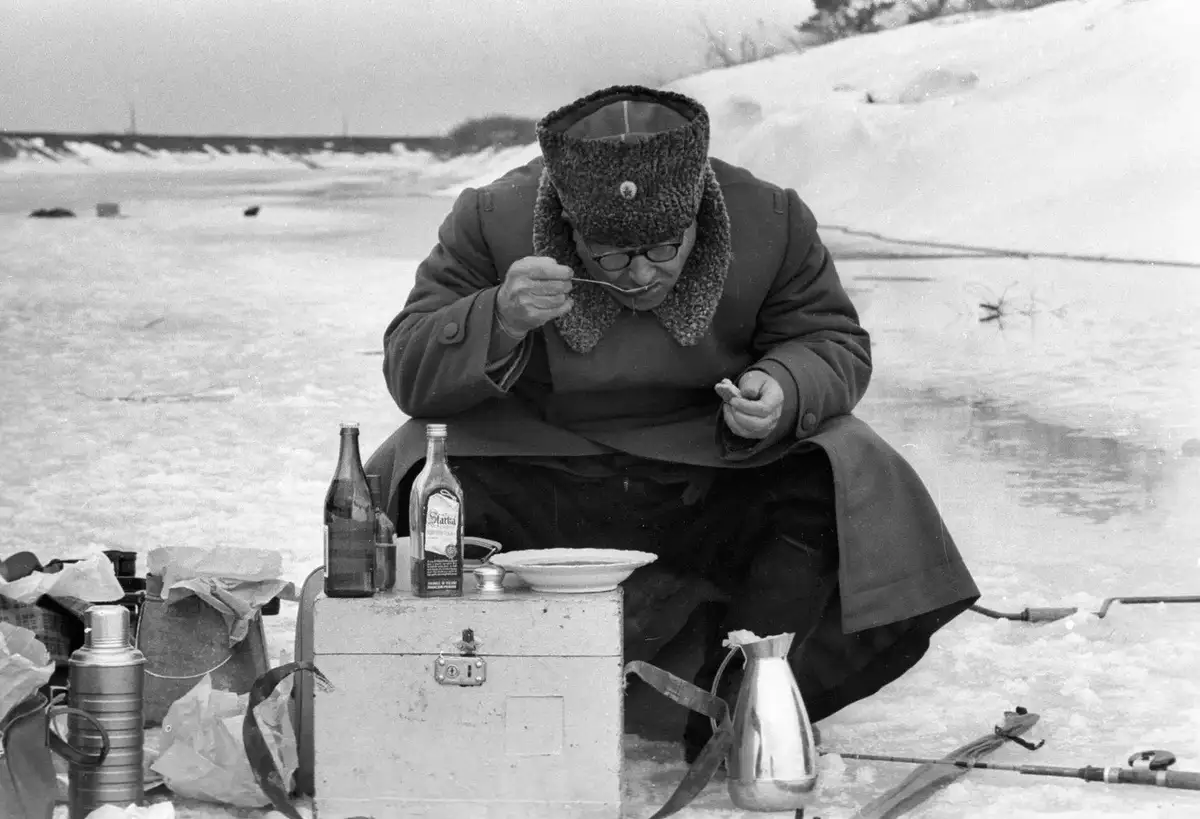 a black-and-white photo shows a man in heavy winter clothing and a fur hat, sitting on ice and eating from a bowl while…