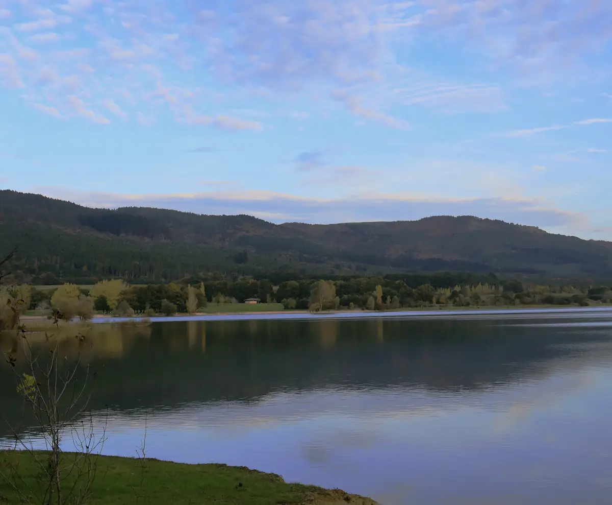 A serene landscape photo of a calm lake reflecting a partly cloudy sky, with forested hills in the background and a grassy…