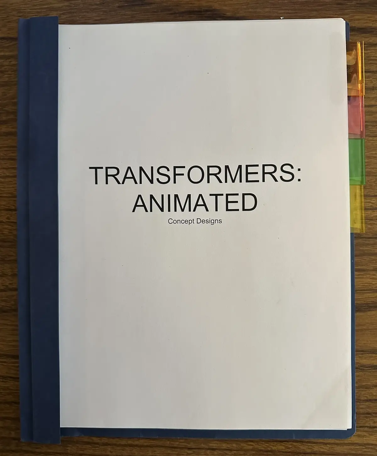 A plain white binder with a blue spine sits on a wooden surface, its cover printed with the title 'TRANSFORMERS: ANIMATED'…