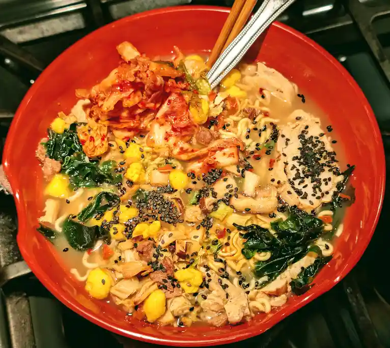 A close-up photo shows a large, red bowl filled with ramen noodles, broth, sliced chicken, kimchi, spinach, corn, mushrooms…