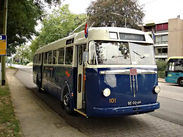 A vintage blue and cream trolleybus, numbered 101 and bearing the license plate BE-00-64, is parked on a paved street with…