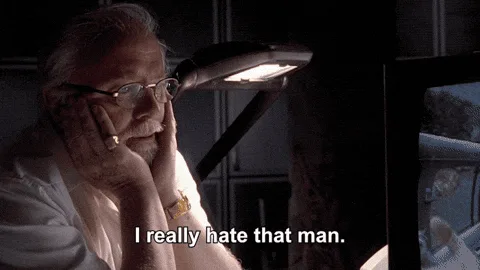 The image shows a man with glasses and a mustache, sitting in a dimly lit room with a desk lamp, resting his chin on his…