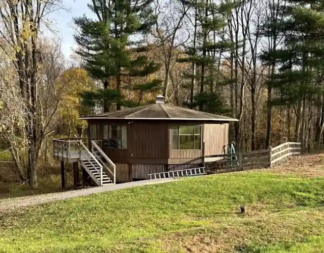 a small wooden cabin with a deck and stairs sits on a grassy slope surrounded by trees, with overlaid text reading 'this is…