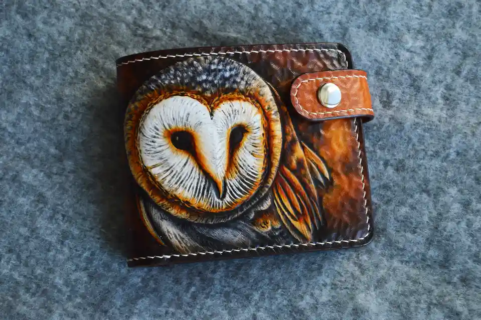 A brown leather wallet with white stitching lies on a gray textured surface, featuring a detailed, hand-painted image of a…