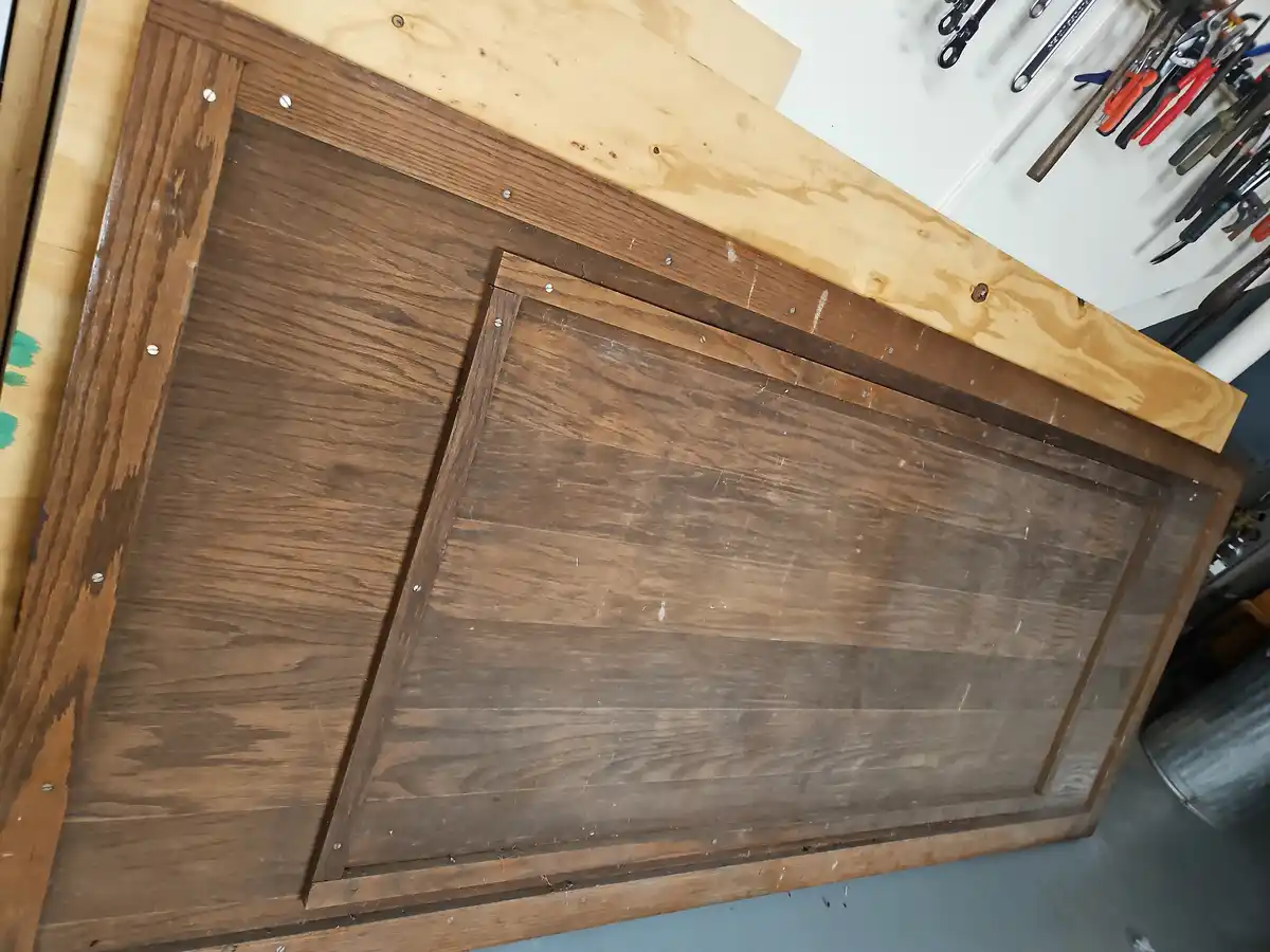 A wooden cabinet door with a dark stain and visible screws lies on a workbench in a workshop, with tools hanging on the wall…