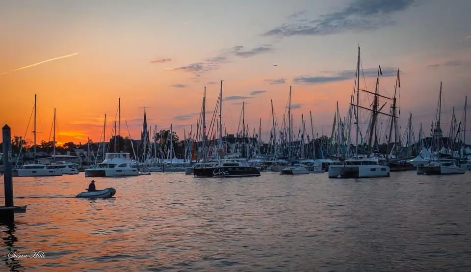 A serene sunset over a harbor filled with numerous sailboats and yachts, their masts silhouetted against the orange and pink…