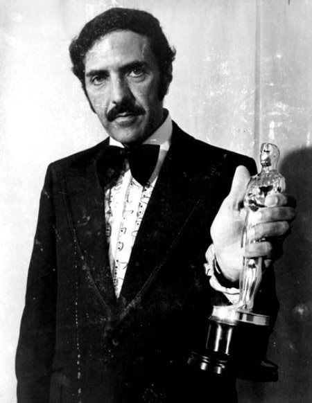 A black-and-white photo shows a man with a mustache in a tuxedo holding an Oscar statuette. He has a serious, slightly smug…