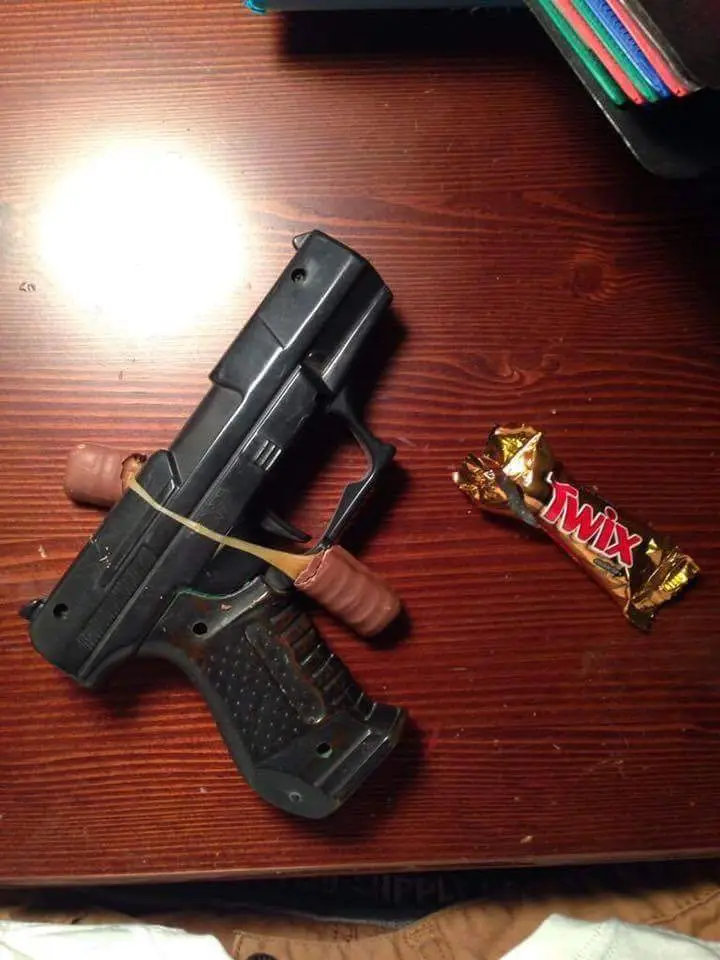 A black toy gun with a rubber band around its grip, holding two brown Twix candy bars, sits on a wooden table next to a…