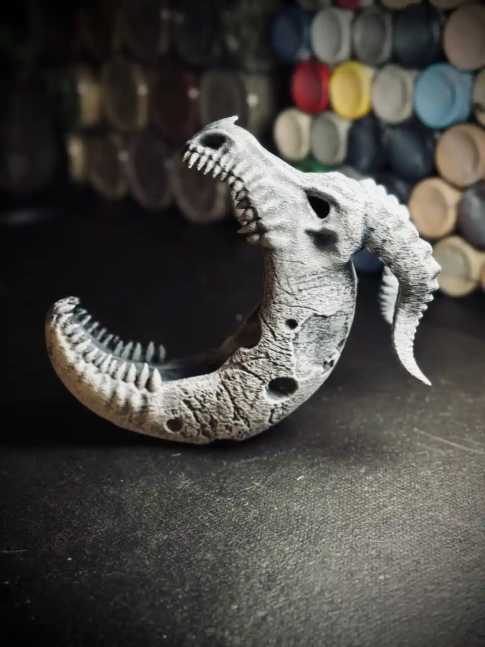 A 3D-printed dinosaur skull model, resembling a theropod with sharp teeth and a curved jaw, is positioned on a dark surface…