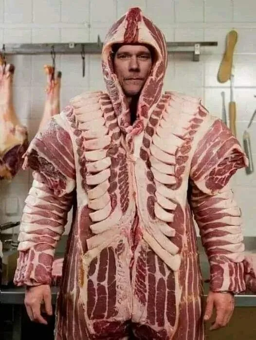 A man stands in a butcher shop wearing a full-body costume made of raw meat, including ribs and fat, with the hood pulled up…