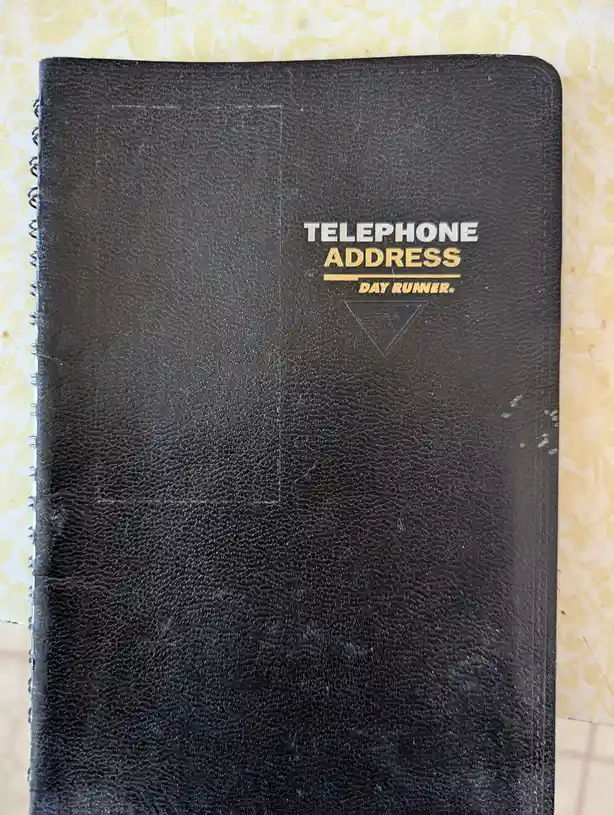 A close-up of a well-worn black spiral-bound Day Runner telephone address book with textured cover, sitting on a yellow…
