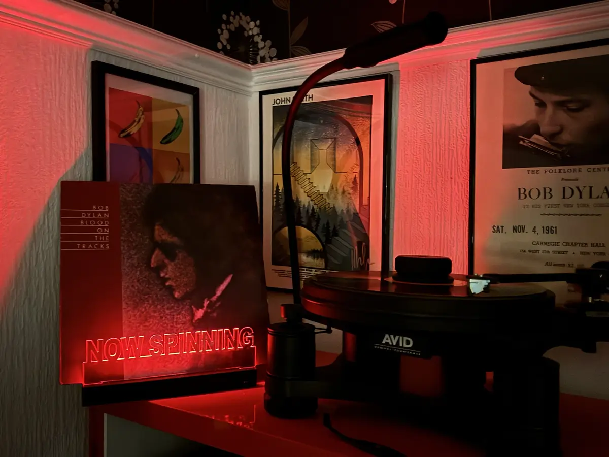 A dimly lit room bathed in red light features a turntable with a vinyl record playing, next to a glowing red neon sign that…